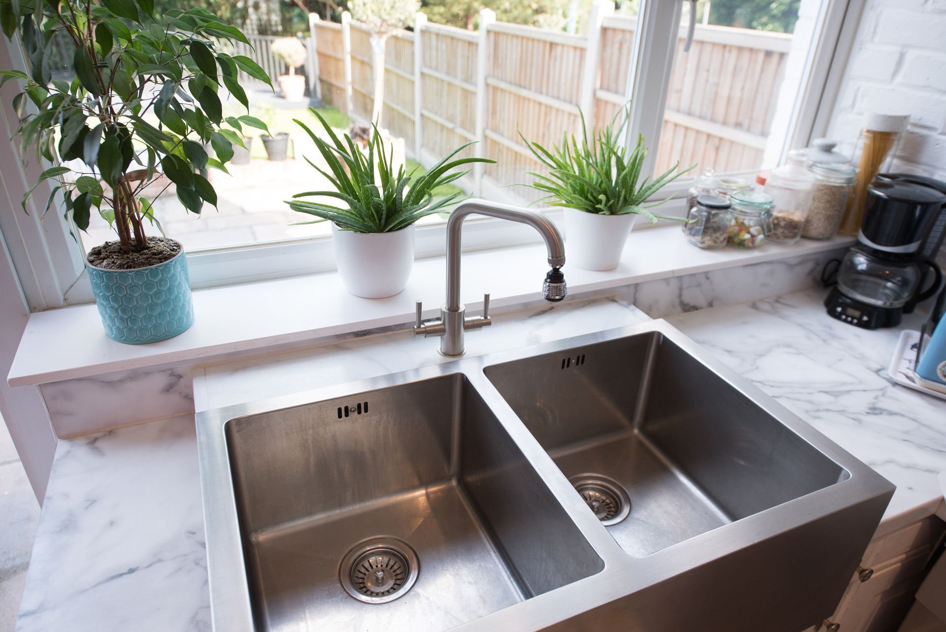 Stainless steel double kitchen sink with a window above, plants on windowsill.