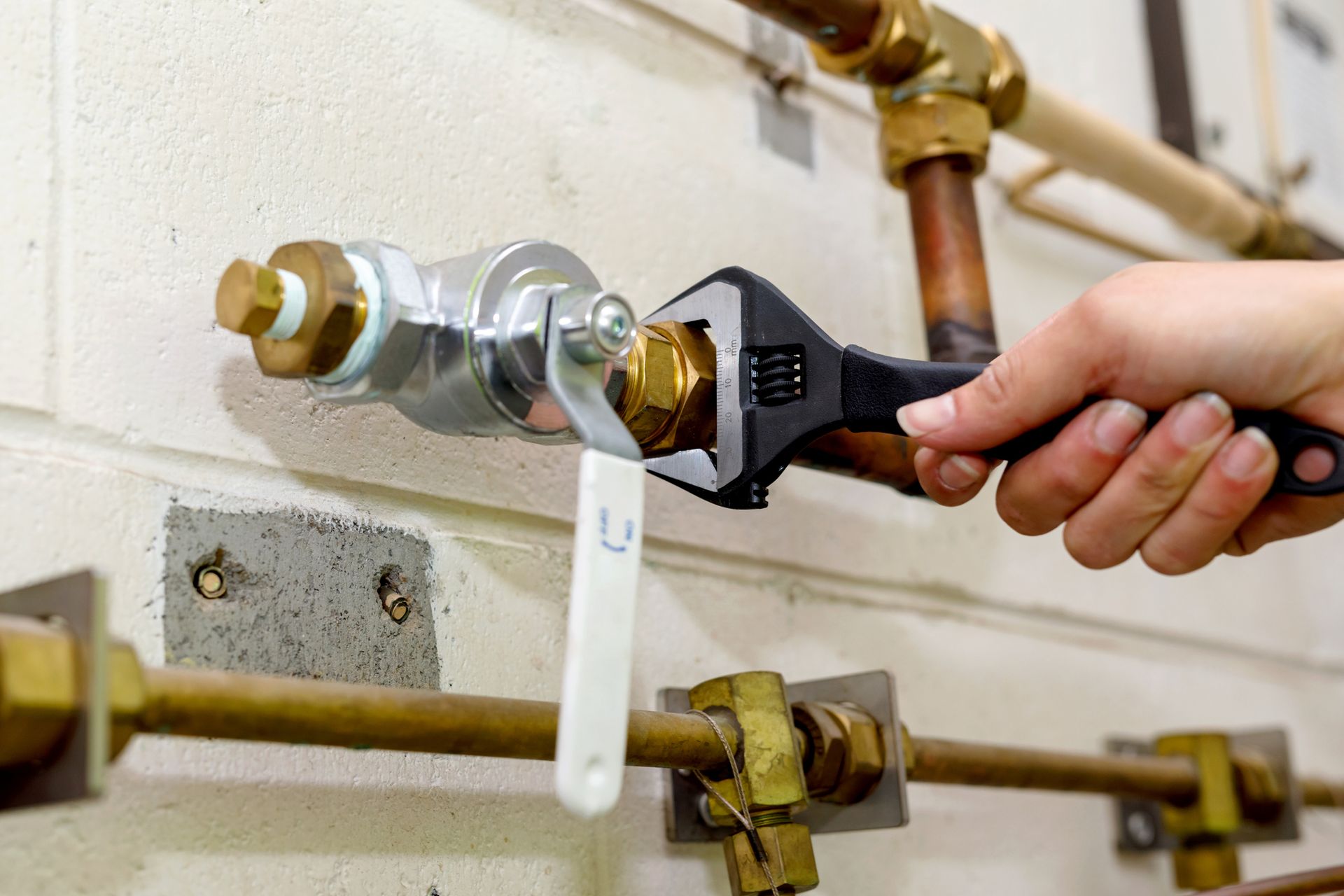 Hand using a wrench to tighten a brass fitting on plumbing against a white wall.