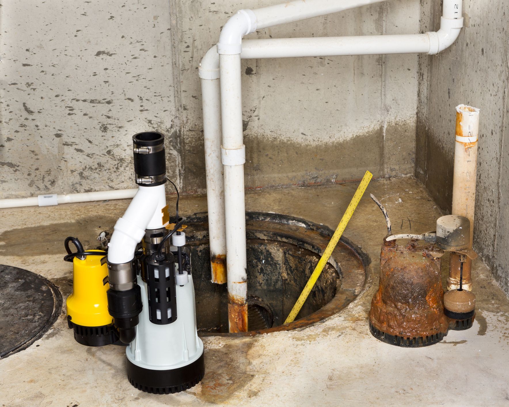 Sump pump system in a basement; a yellow pump next to the main pump and drain pipes.