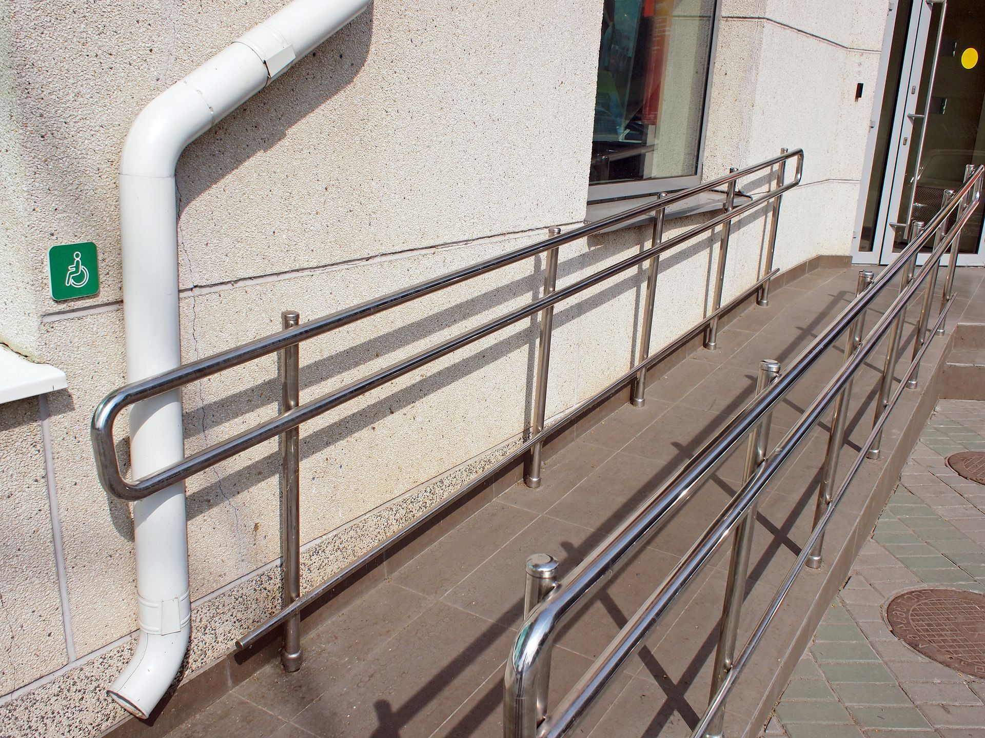Ramp with metal handrails for wheelchair access at a building entrance, green disability sign on the wall.