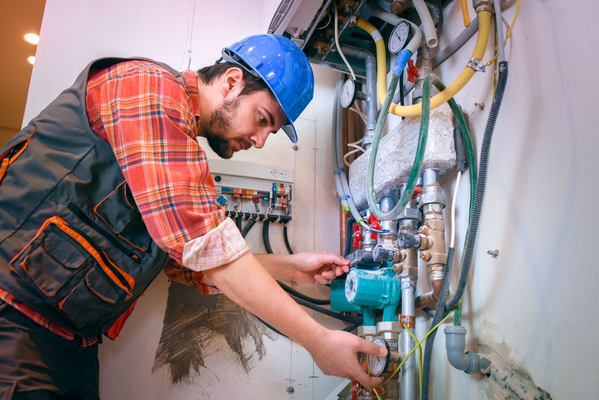 A technician in a blue hard hat and plaid shirt repairs a water heater.