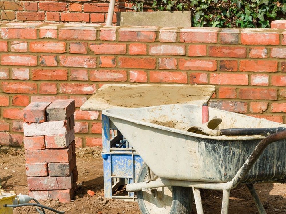 A wheelbarrow with mortar sits beside a stack of bricks and a yellow power tool in front of a red brick wall.