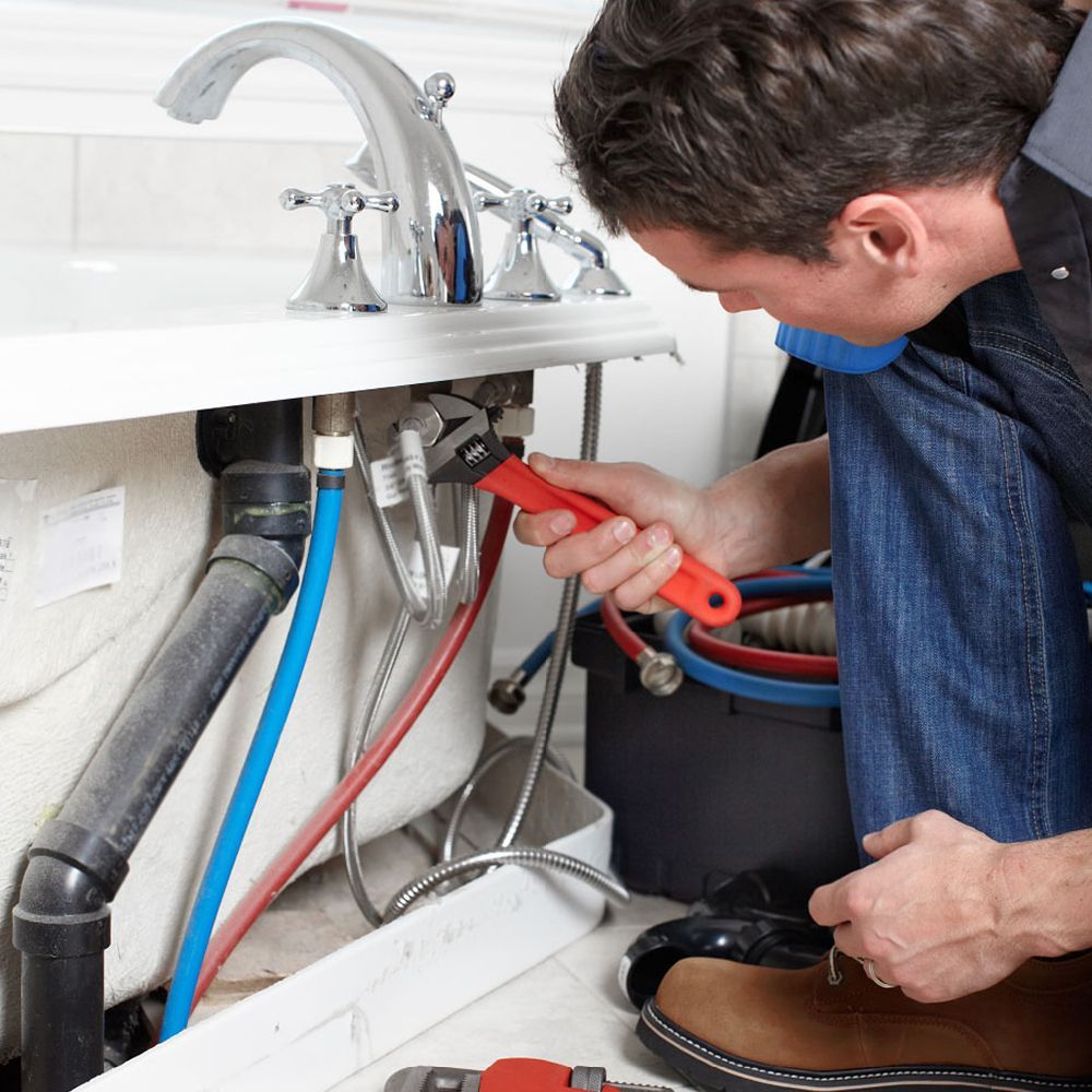 A plumber uses a red wrench to tighten connections beneath a bathtub faucet.