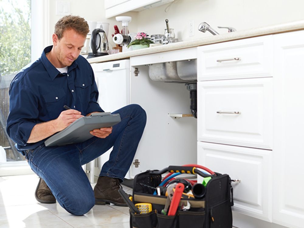 A plumber in a blue shirt kneels in a kitchen, writing on a clipboard near a sink with a tool bag on the floor.