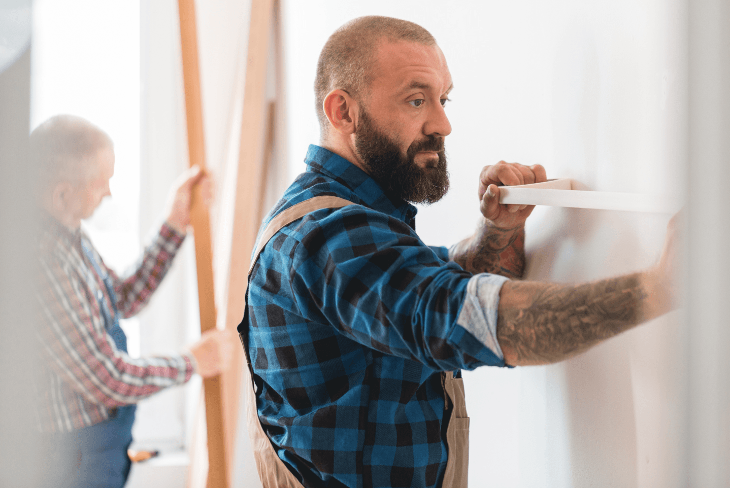 A person in a blue plaid shirt and overalls measures a white wall while another person works with lumber in the background.