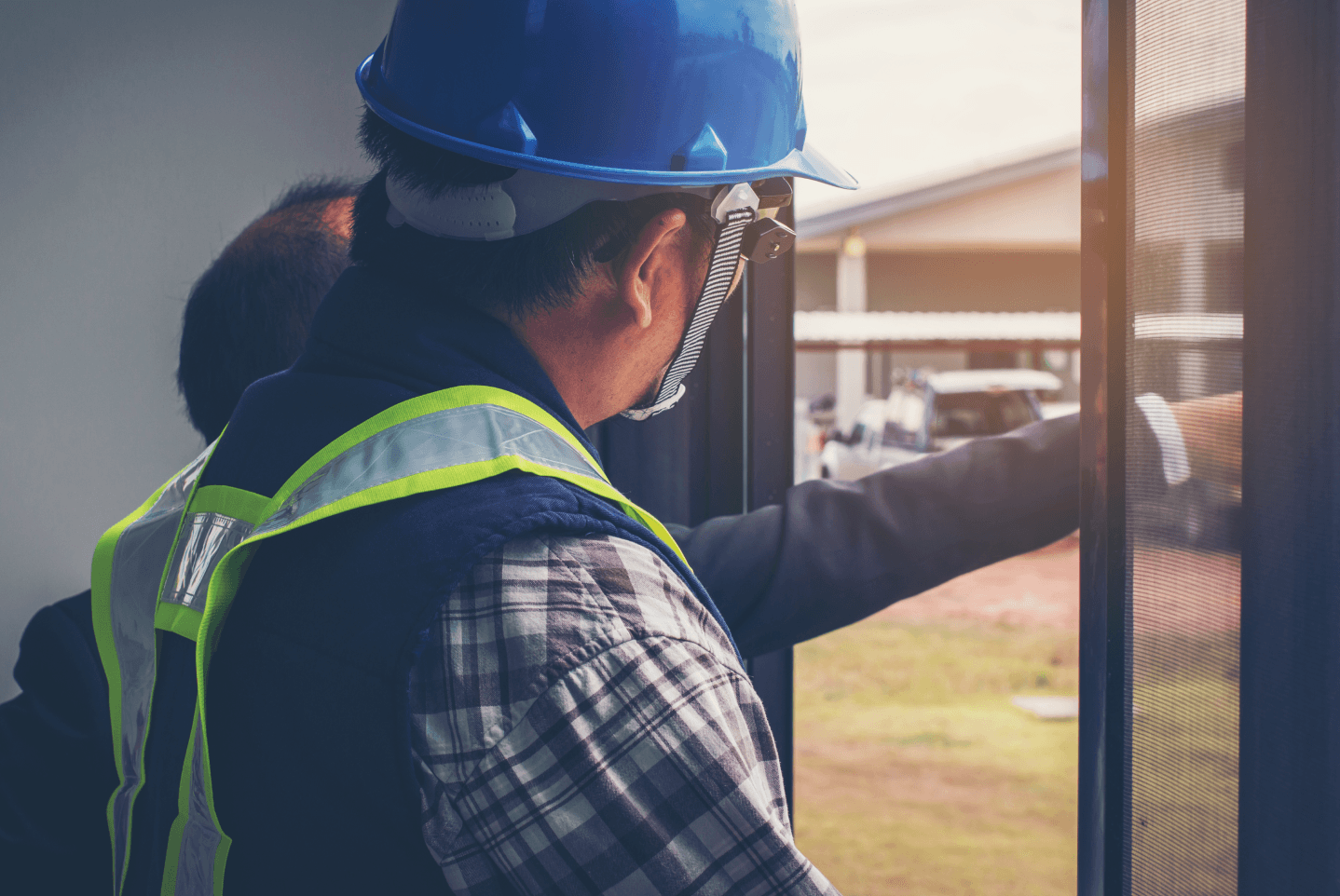 A person wearing a blue hard hat and high-visibility vest stands in a doorway, gesturing toward an outdoor construction site.