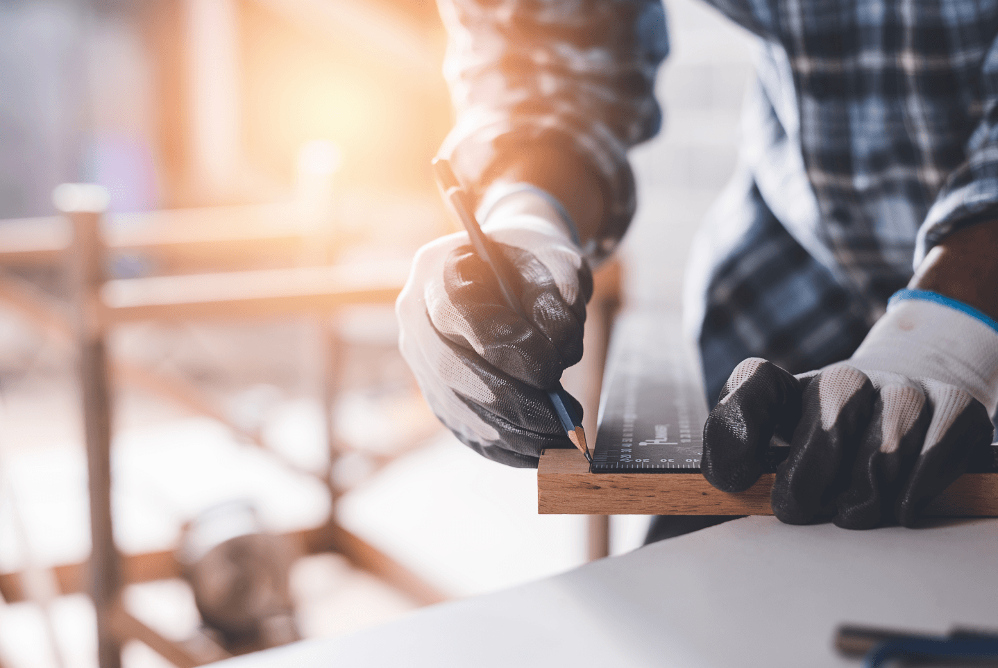 A person wearing work gloves marks a line on a wooden board with a pencil and a metal square in a workshop setting.