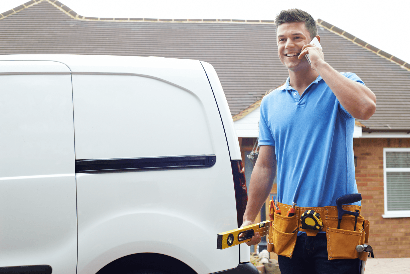 A tradesperson wearing a tool belt and holding a level walks by a white van while talking on a smartphone.