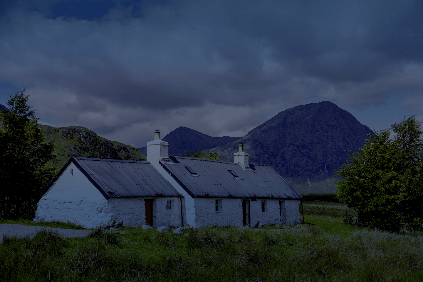 A small, white cottage with a dark roof sits in a grassy valley before rugged mountains under a cloudy sky.