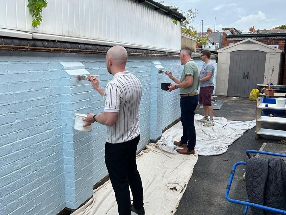 Three people in casual clothing painting a brick wall light blue, working together on a drop cloth in an outdoor area.