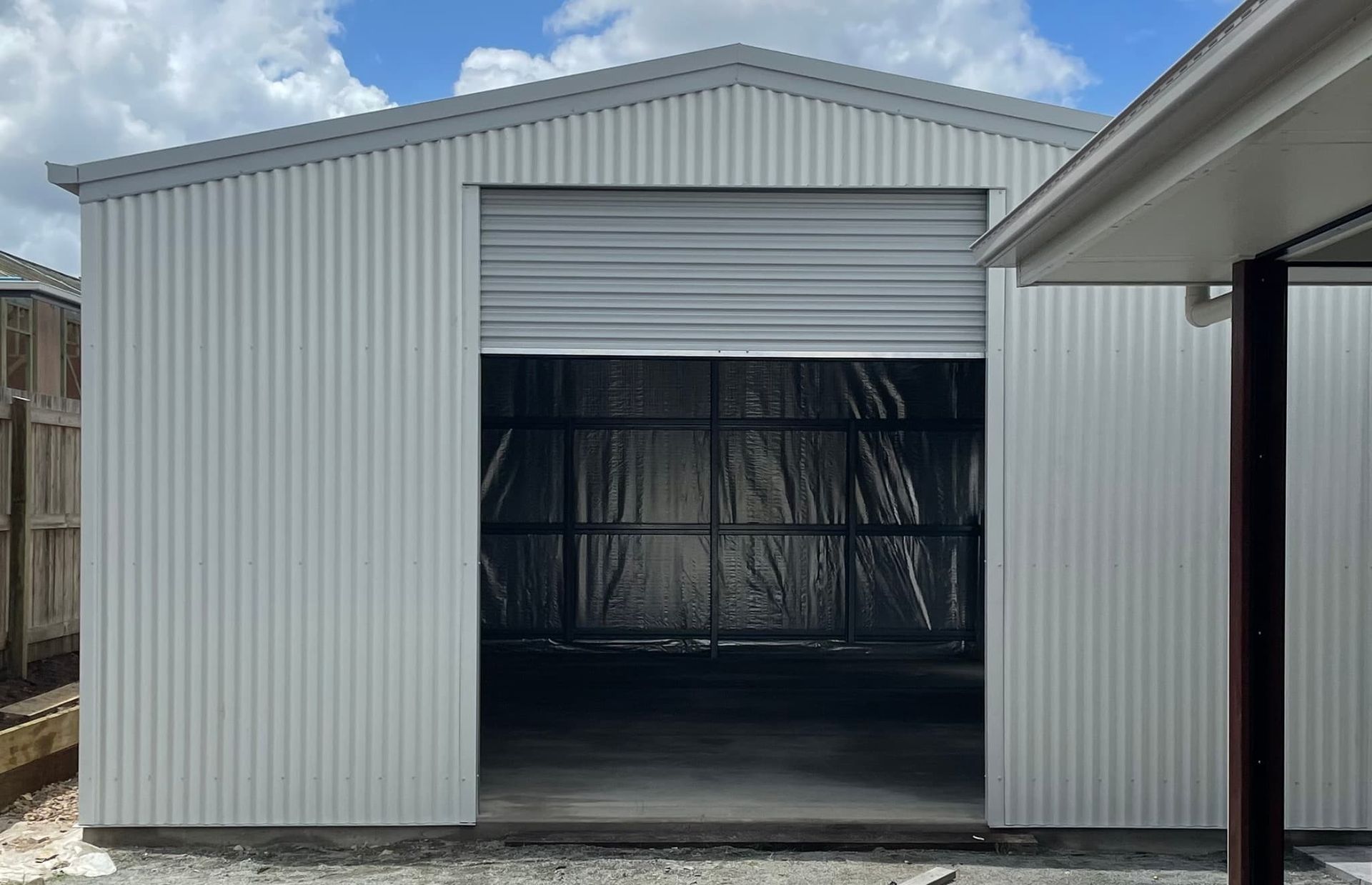A white garage with a roller door is sitting next to a house. — Just Sheds In Ipswich, QLD