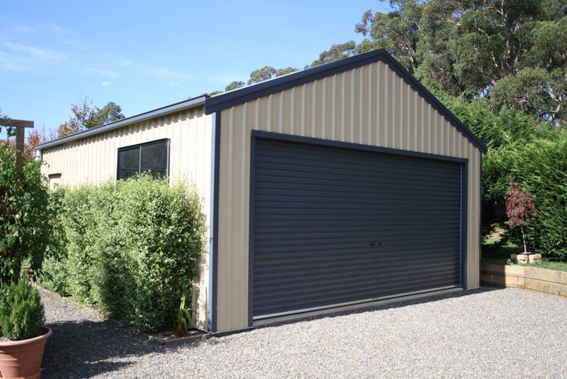 A Garage With a Black Garage Door is Surrounded by Trees and Bushes — Just Sheds In Kunda Park, QLD
