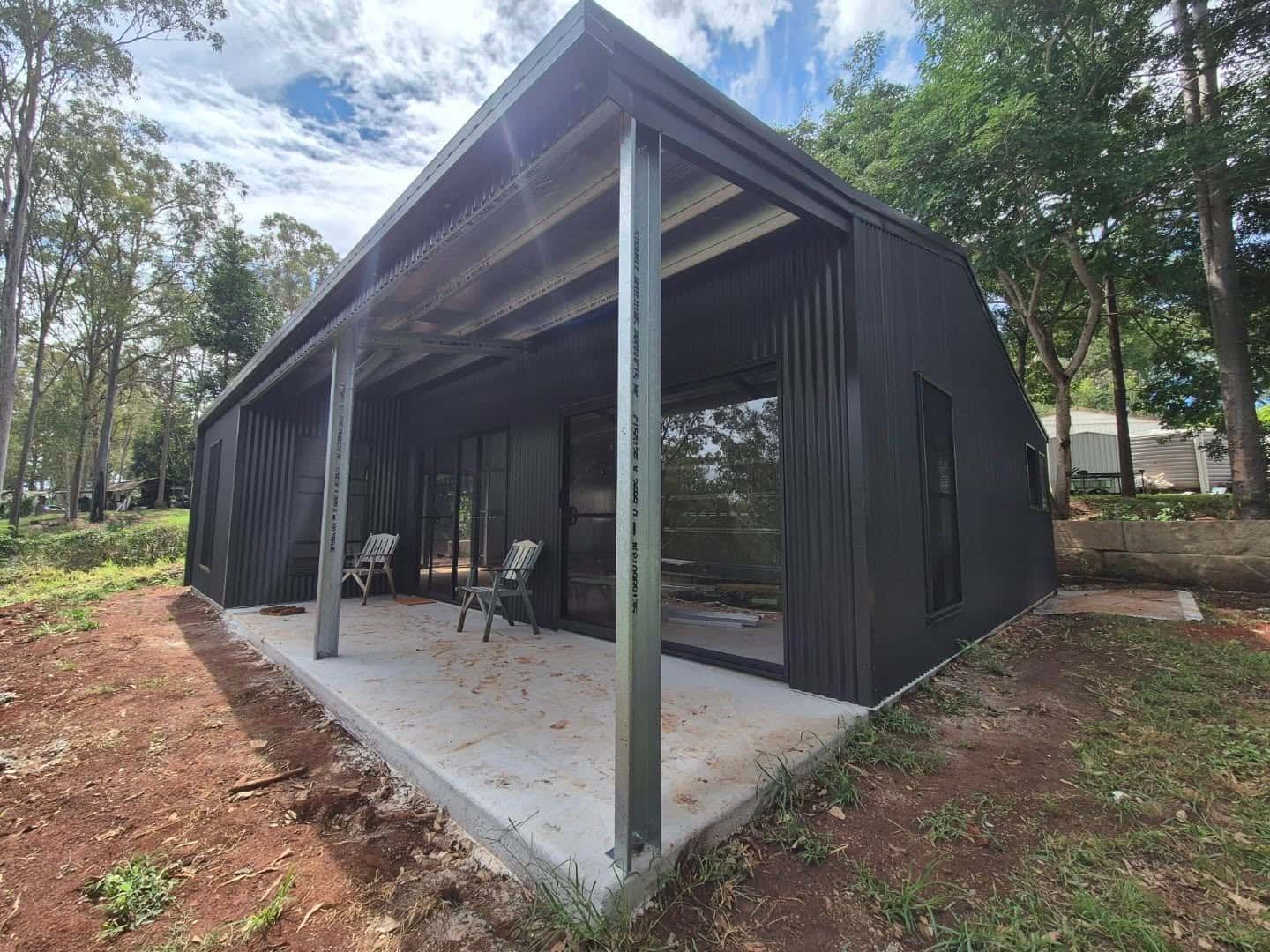 A small black house with a porch and chairs in front of it. — Just Sheds In Sunshine Coast, QLD