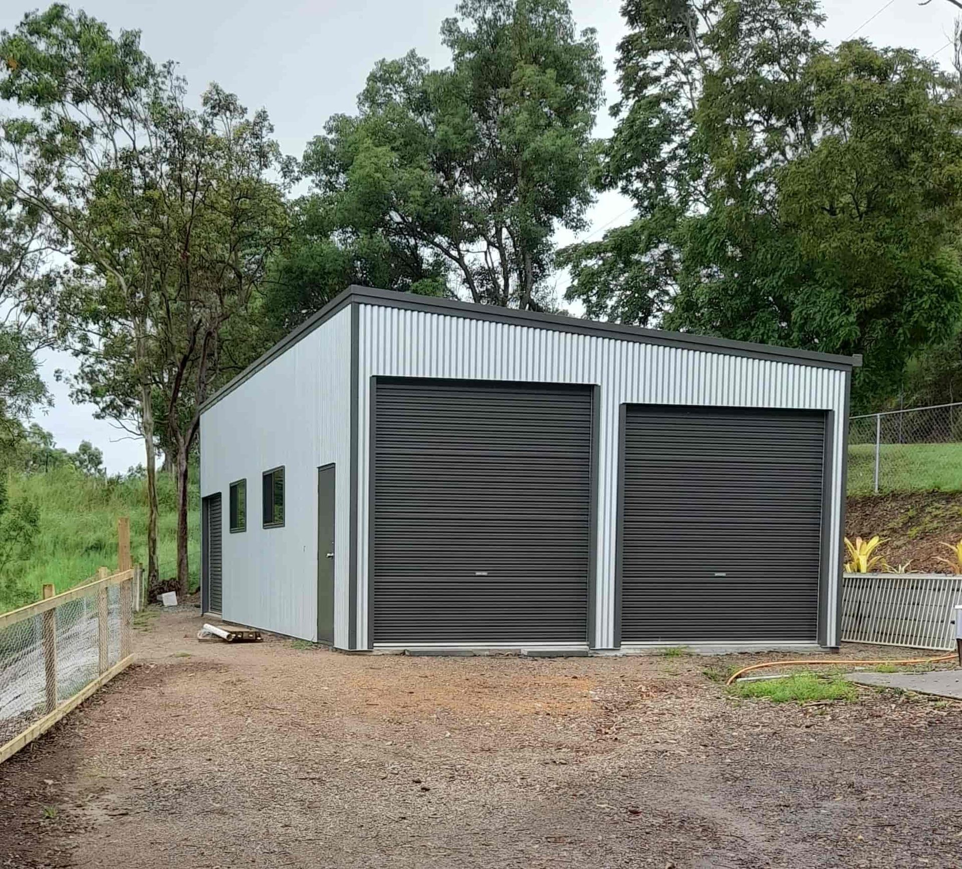 A garage with two black garage doors is surrounded by trees — Just Sheds In Kunda Park, QLD