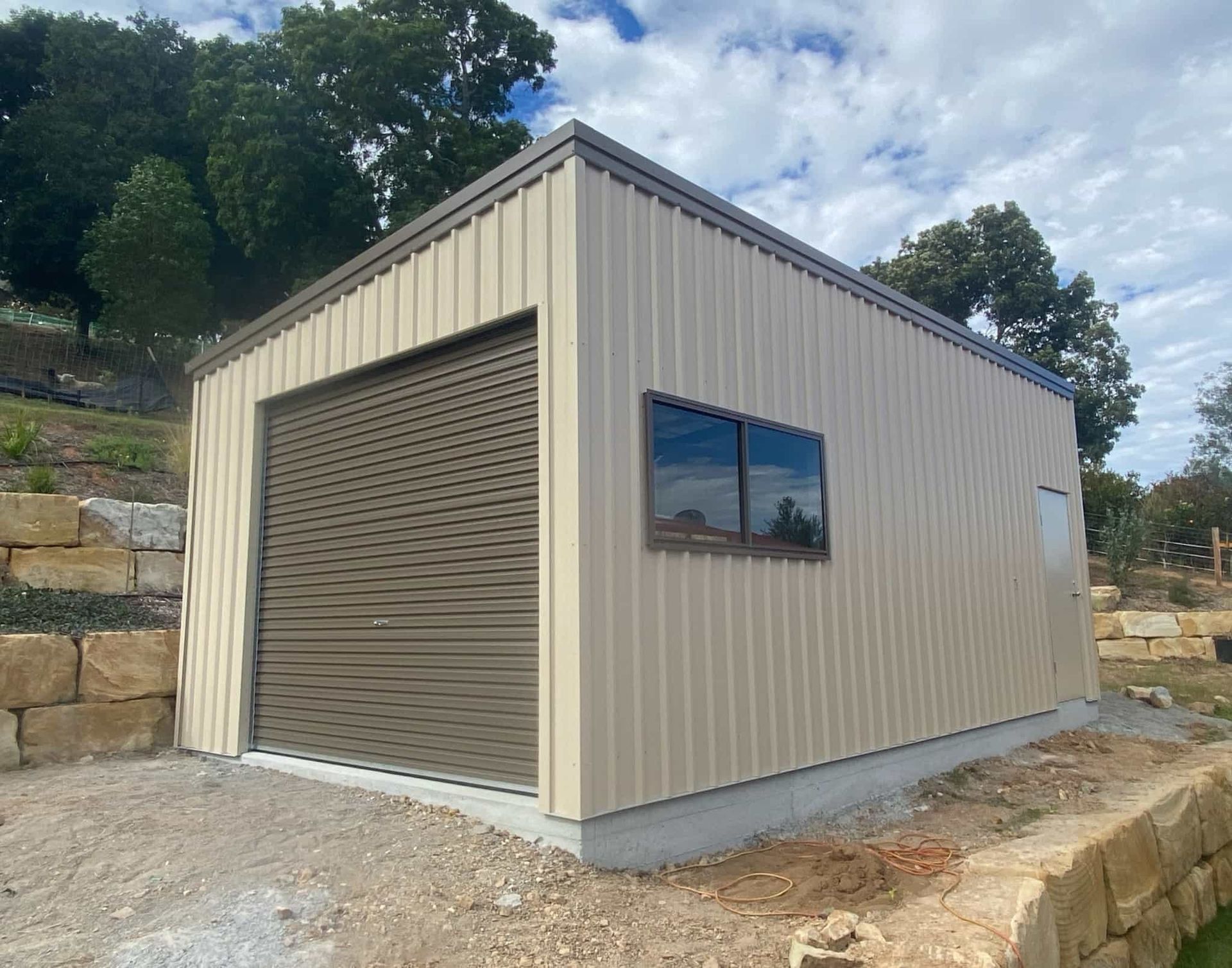 A garage with a roller door and a window is sitting on top of a dirt hill. — Just Sheds In Brendale, QLD