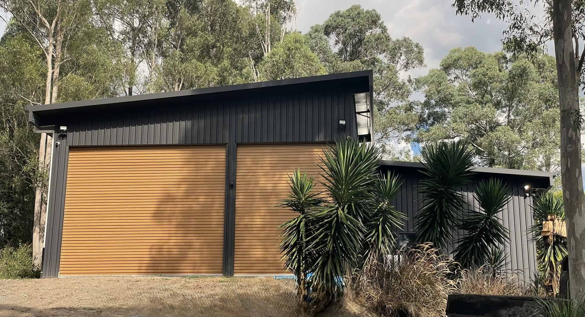 A small house with a wooden garage door is surrounded by trees. — Just Sheds In Sunshine Coast, QLD