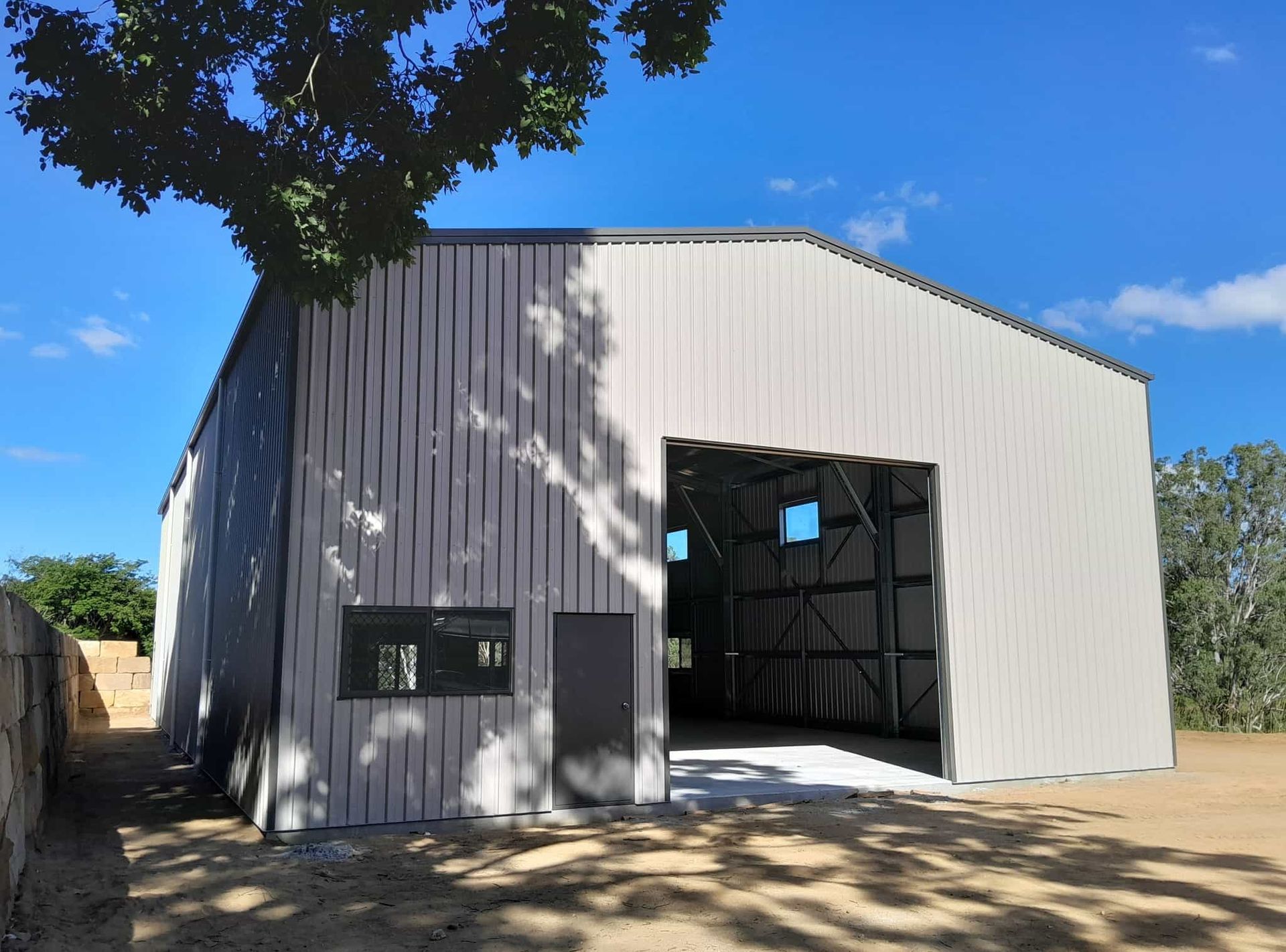 A large metal building with a large door is sitting in the middle of a dirt field. — Just Sheds In Gympie, QLD
