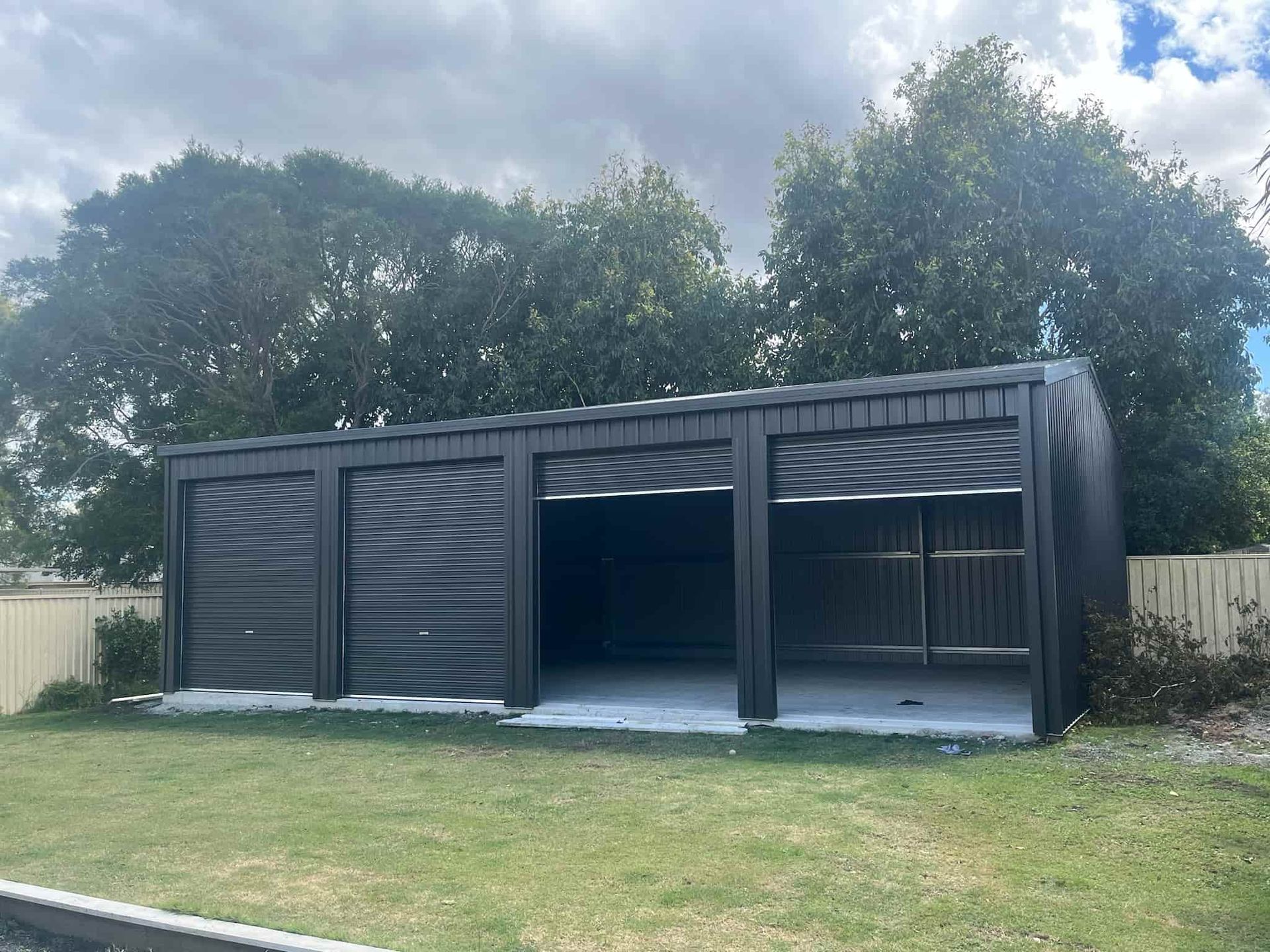 A large black garage is sitting in the middle of a lush green yard. — Just Sheds In Kunda Park, QLD