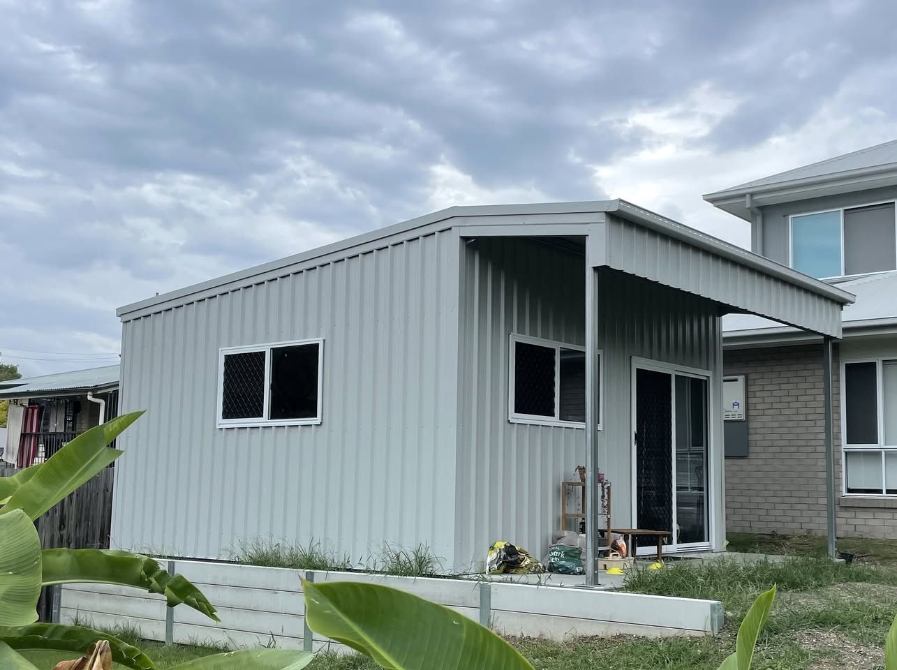 A small white building with a porch next to a house. — Just Sheds In Kunda Park, QLD