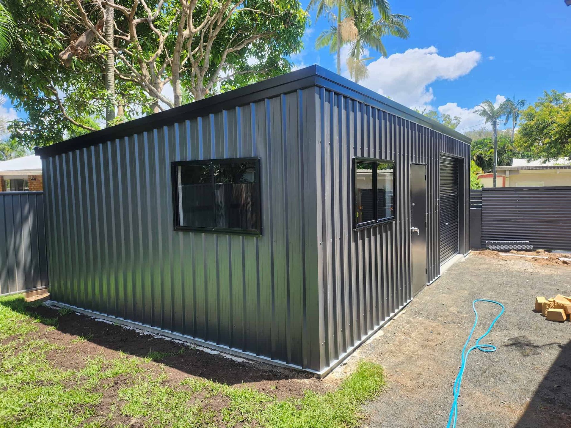 A black metal building with a fence in the background and a dog standing in front of it. — Just Sheds In Kunda Park, QLD