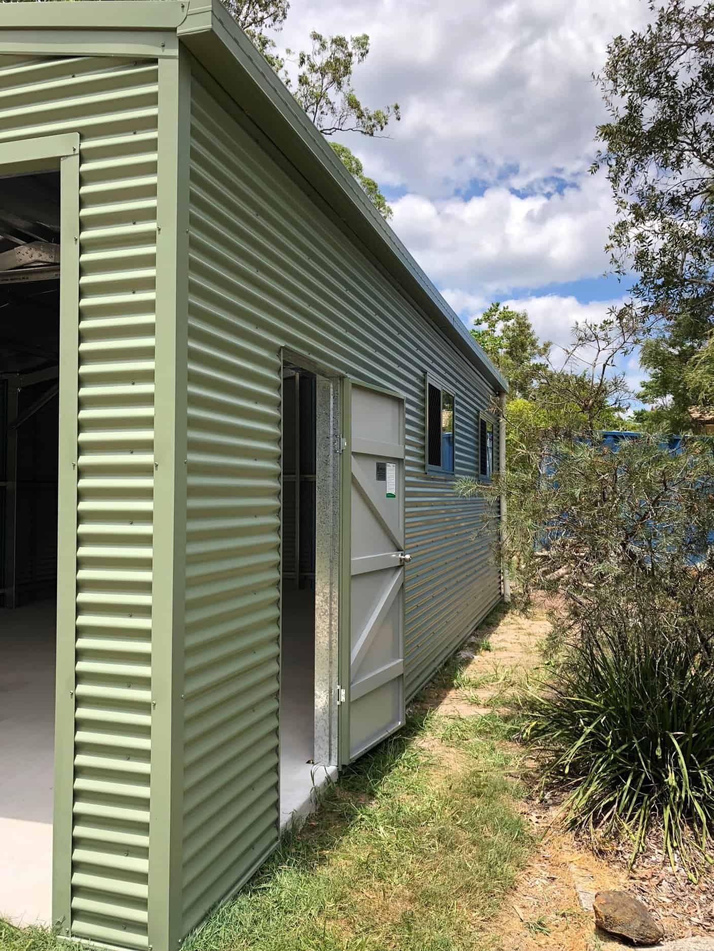 A green shed with a white door is sitting in the grass. — Just Sheds In Gympie, QLD