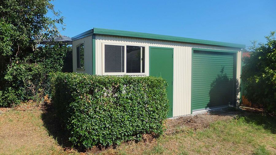 A Green and White Shed With a Green Door is Surrounded by Trees and Bushes — Just Sheds In Kunda Park, QLD