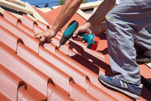 A man is working on a roof with a drill. Residential roofing contractor inspecting shingles on sloped roof under blue sky.