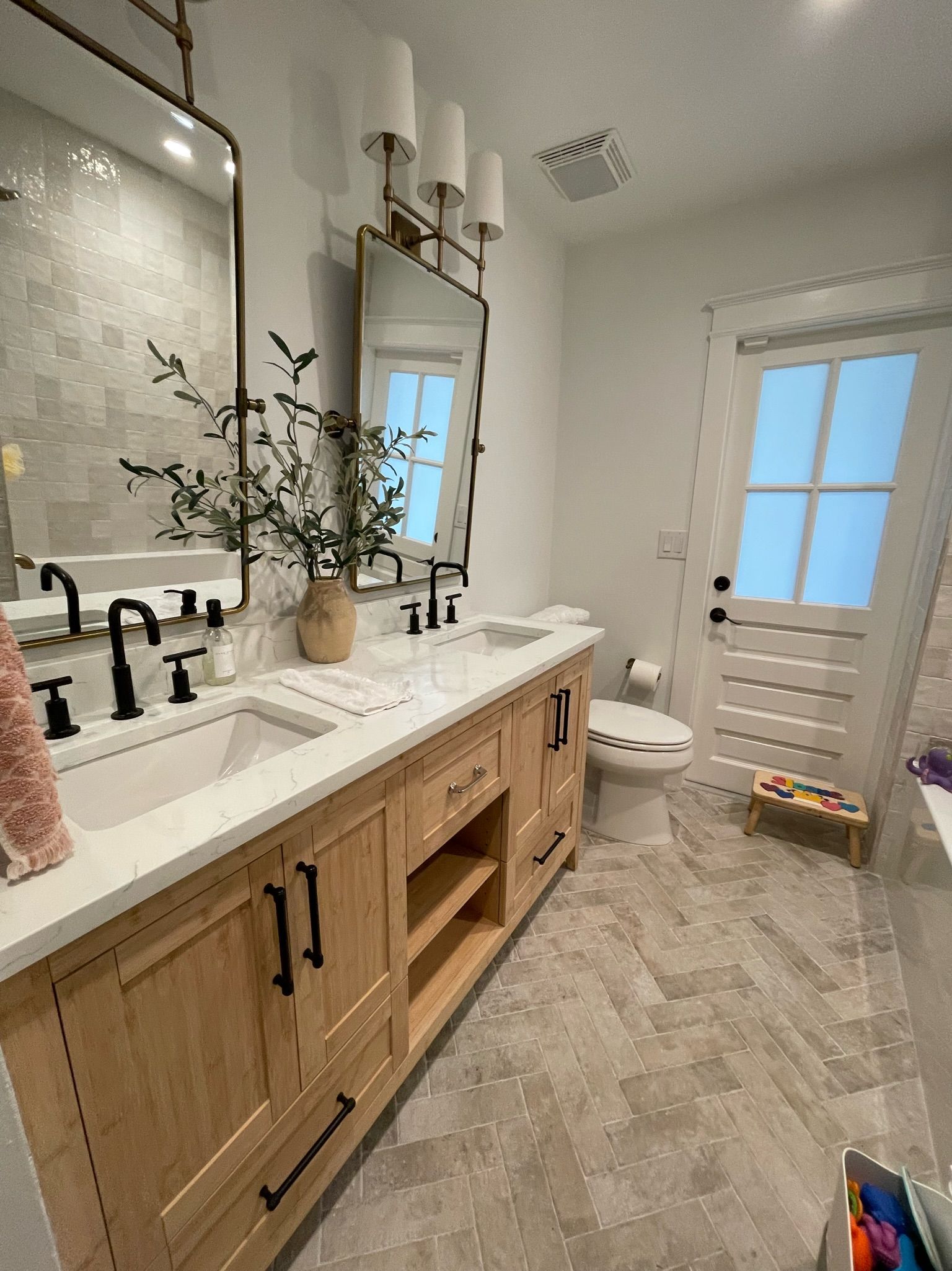 Bright bathroom with double wood vanity, arched mirror, and tiled floor; window and shower visible.