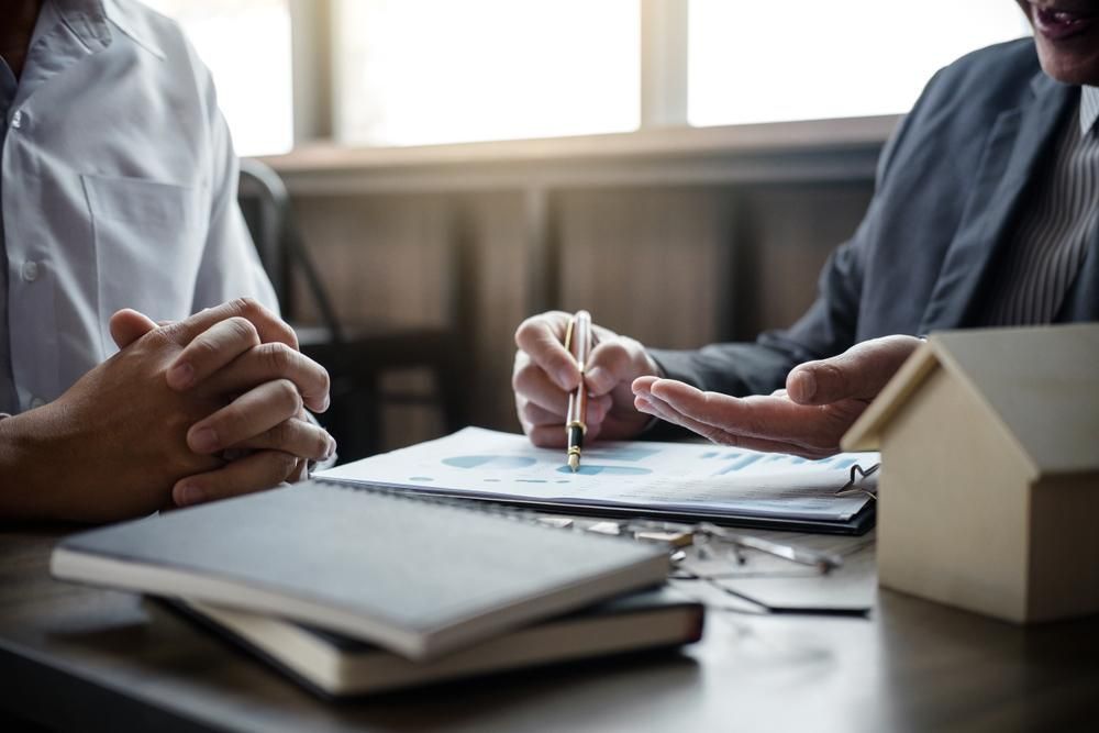 Two Men Are Sitting At A Table With A Model House And A Pen — Howells & Co Legal & Conveyancing In Cessnock, NSW
