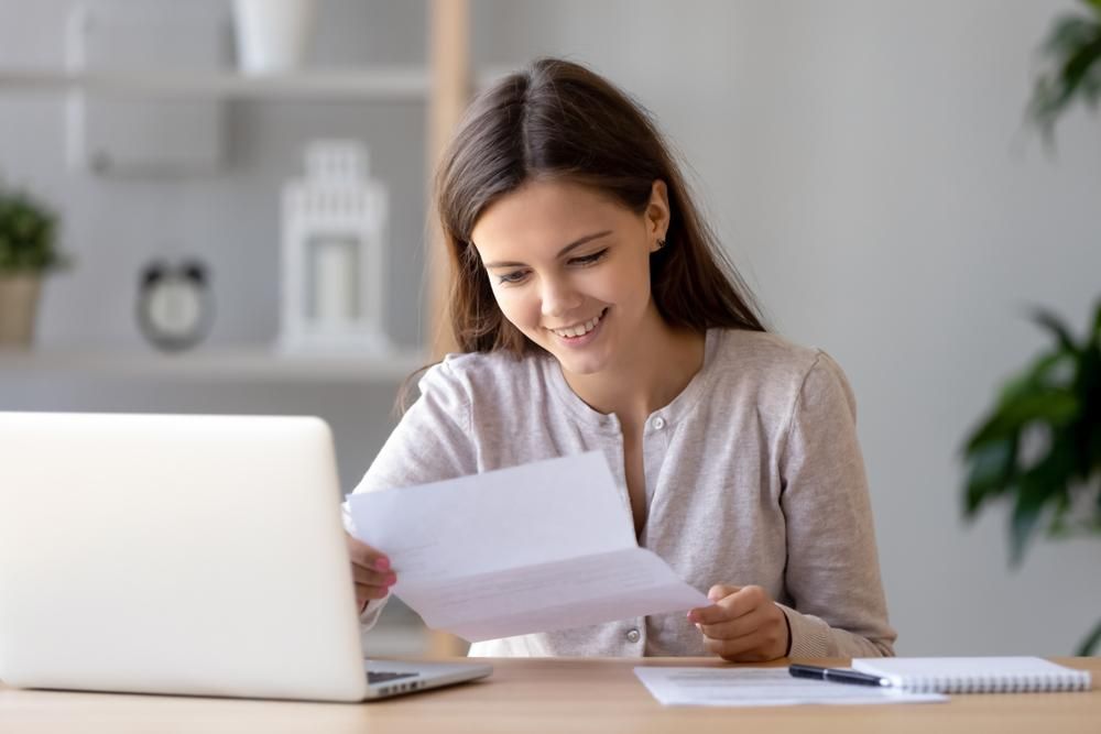 A Woman Is Sitting At A Desk Looking At A Piece Of Paper In Front Of A Laptop Computer — Howells & Co Legal & Conveyancing In Muswellbrook, NSW
