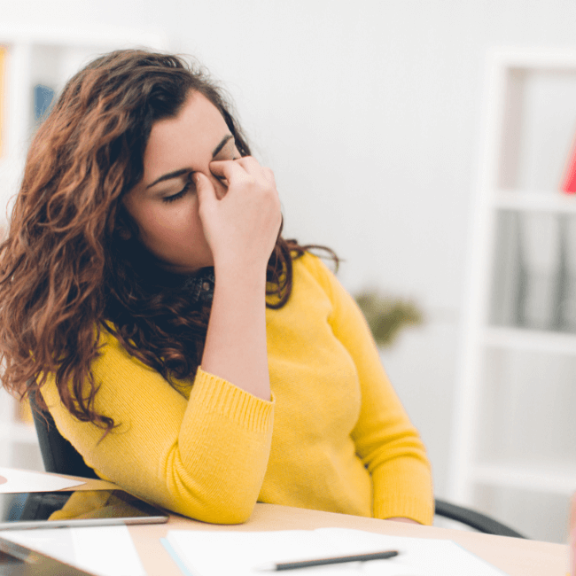 A Woman In A Yellow Sweater Is Sitting At A Desk With Her Hand On Her Face — Howells & Co Legal & Conveyancing In Lake Cessnock, NSW