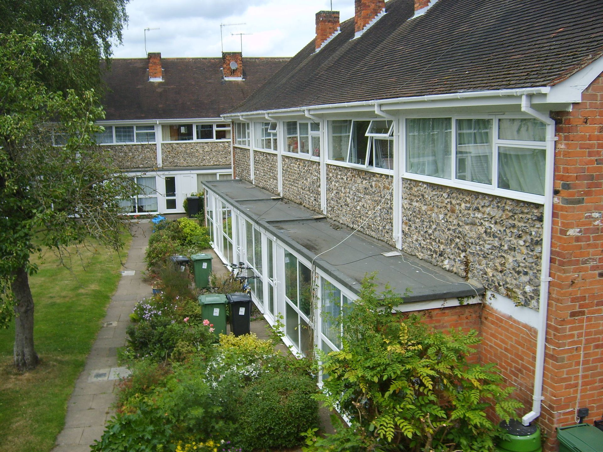 Row of brick houses with a flat-roofed extension in front, a pathway, and greenery.