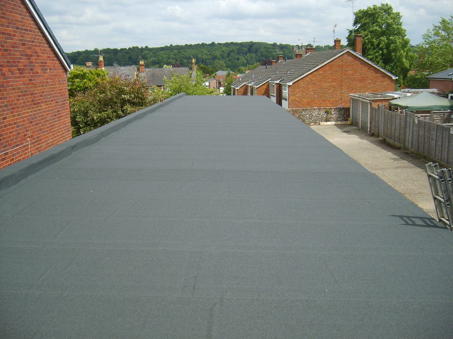 Flat, dark gray rooftop with houses and trees visible in the background under a sunny sky.