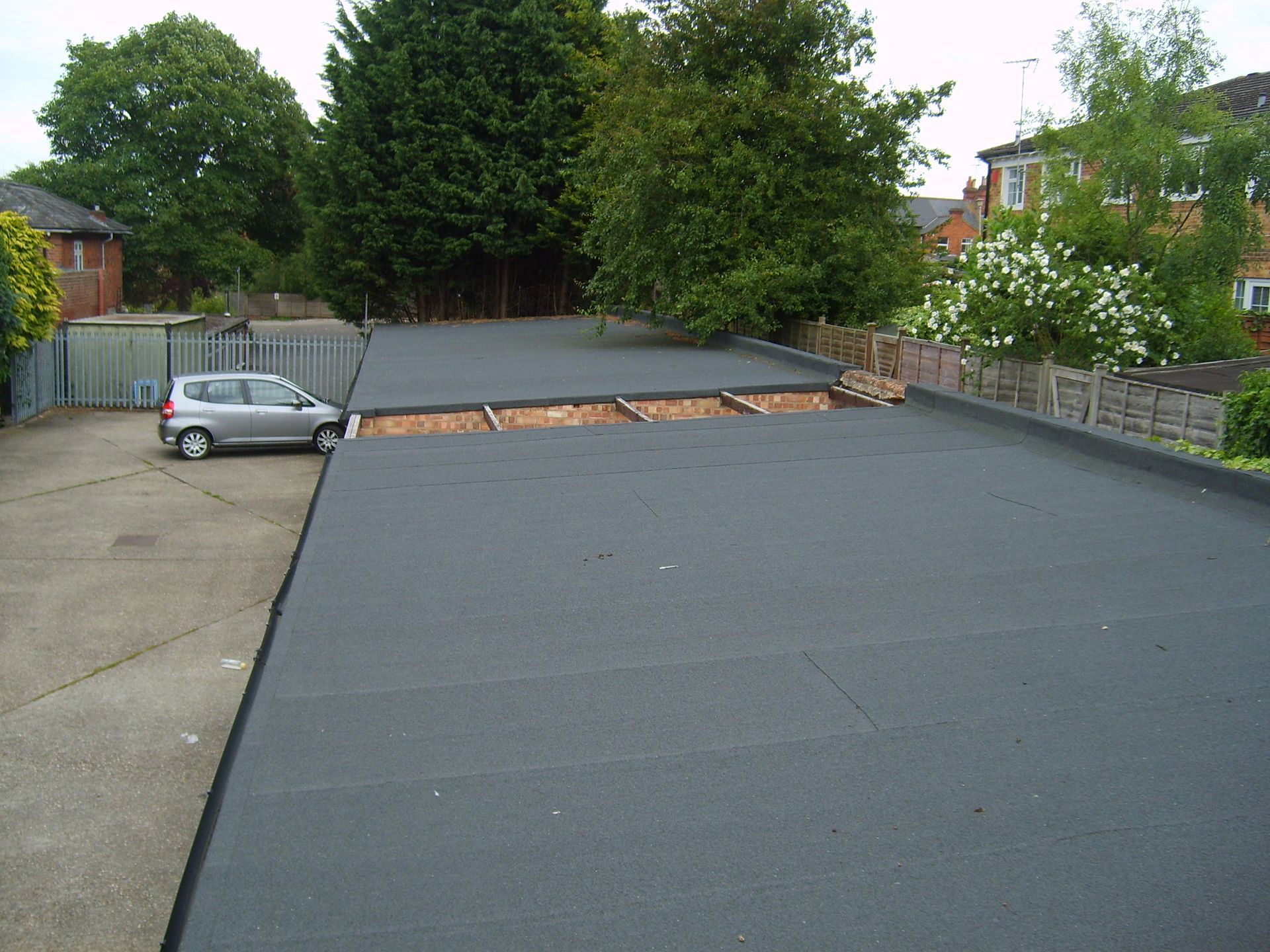 A flat, dark-gray roof with a partially constructed section, parked car, trees, and residential buildings in the background.