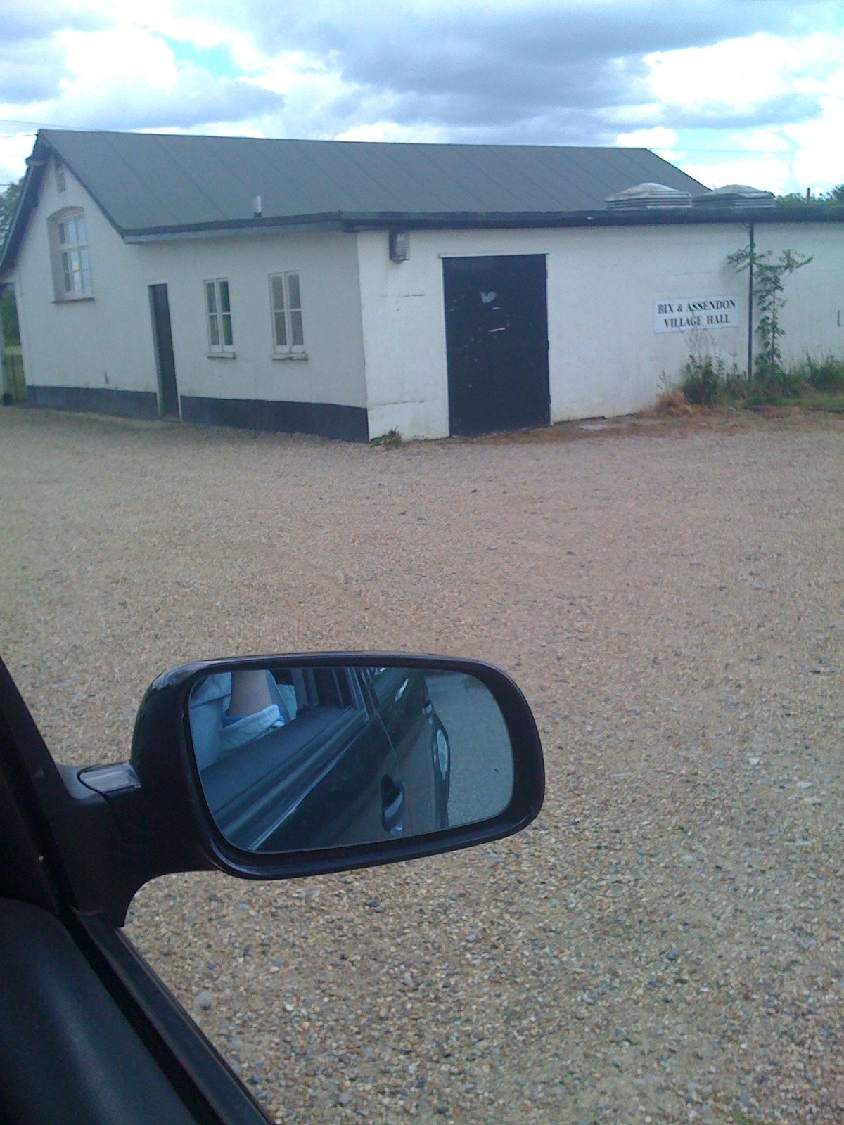 White building with dark door, gravel parking lot, car side mirror reflecting the road. Cloudy sky.