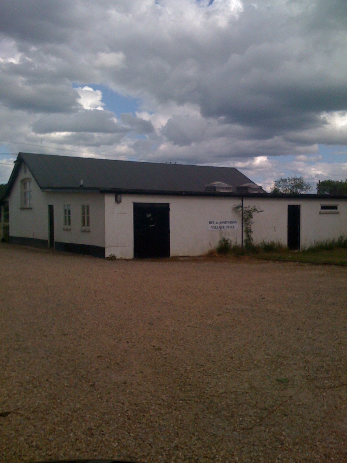 White, single-story building with a dark roof under a cloudy sky. A gravel lot is in the foreground.