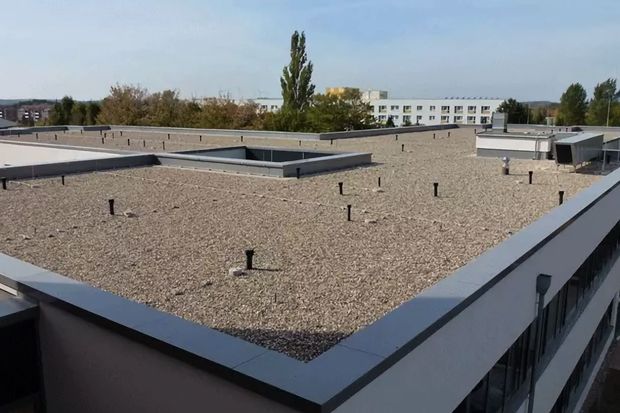 Flat rooftop covered in gravel, with black pipes and an adjacent building in the background under a blue sky.
