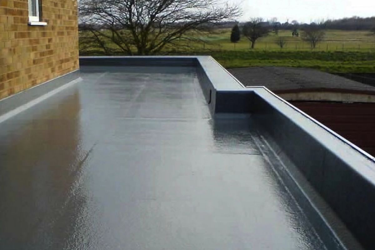 Flat, gray, reflective roof surface with a brick wall and distant green fields visible.