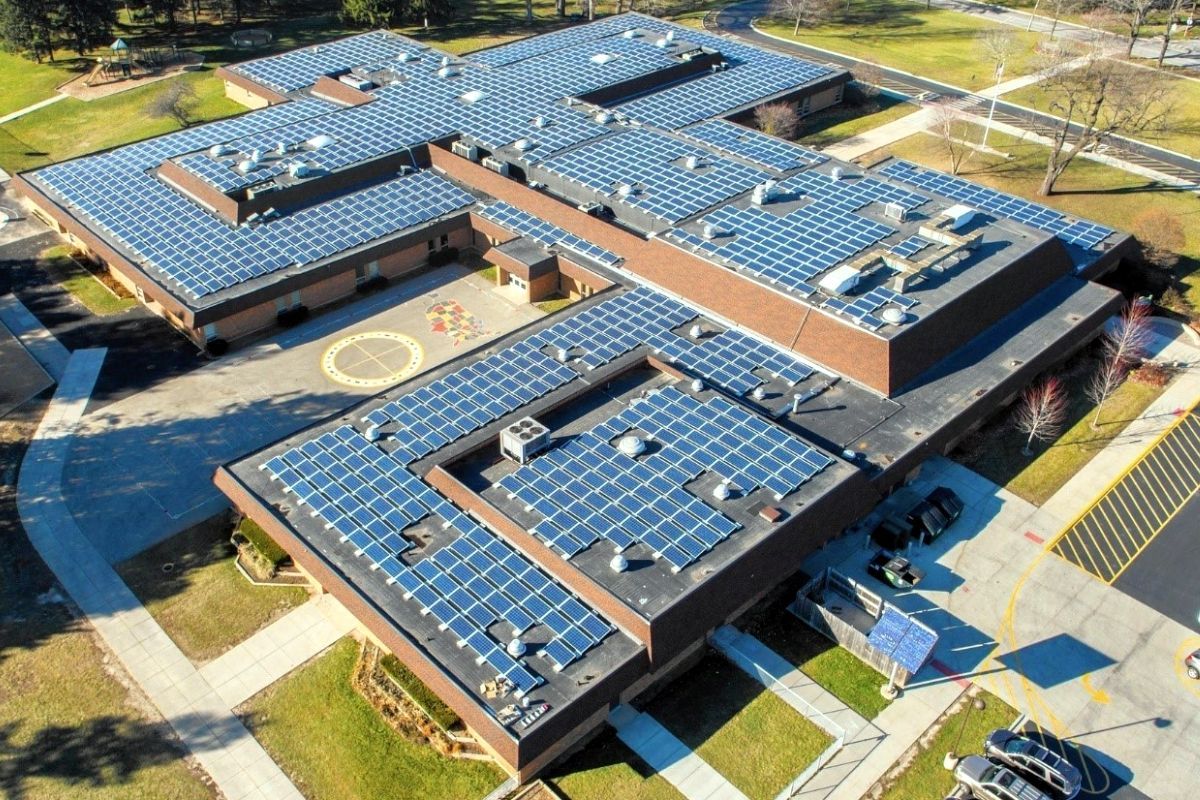 Aerial view of a school building covered in solar panels, surrounded by grassy areas and a parking lot.