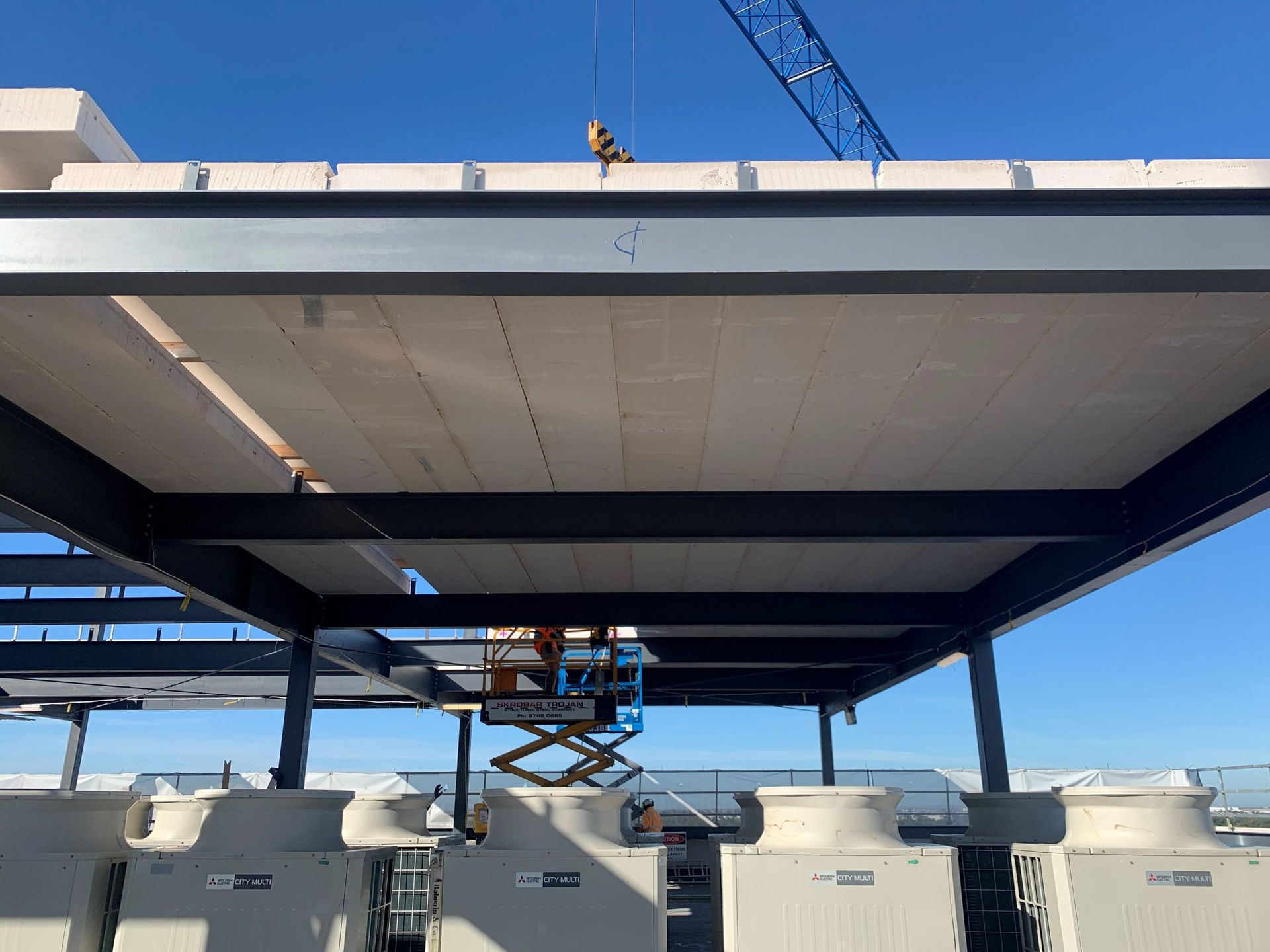 Construction workers installing panels on a steel-framed rooftop structure with a crane. Blue sky visible.