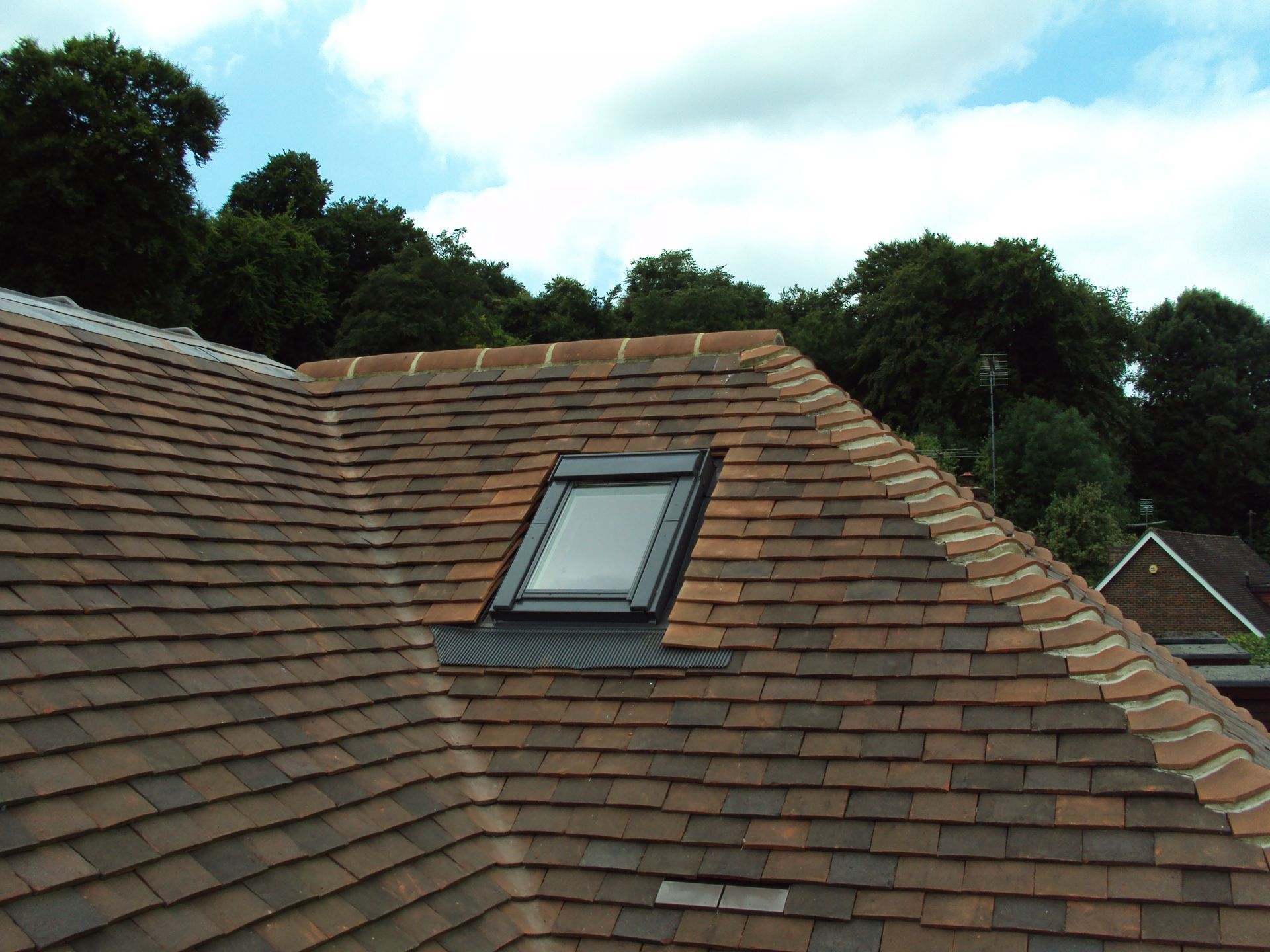 Rooftop with a skylight, covered in brown tiles, against a blue sky and trees.