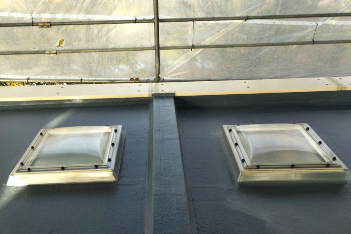 Two skylights on a flat, gray roof, with a gray support beam. Above, a wall of rectangular panels is visible.