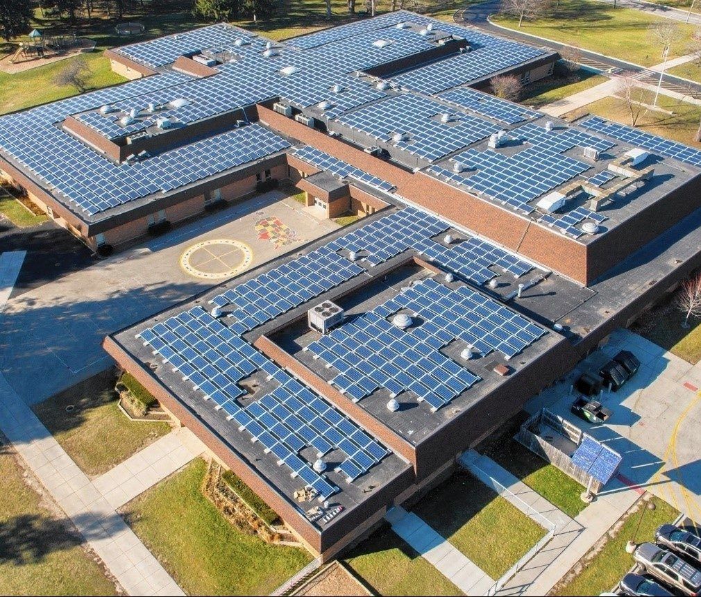 An aerial view of a school building with numerous solar panels on the roof, surrounded by green grass and a parking lot.