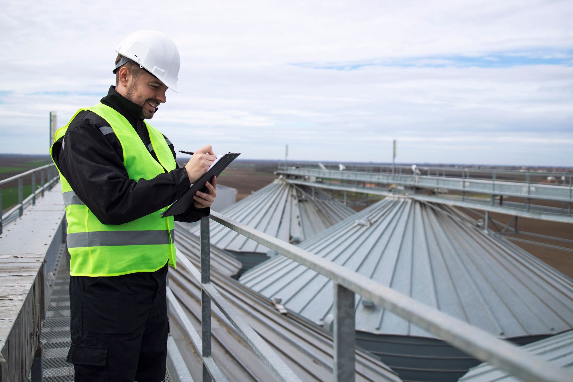 Man in safety gear writing on a clipboard while inspecting on a rooftop.