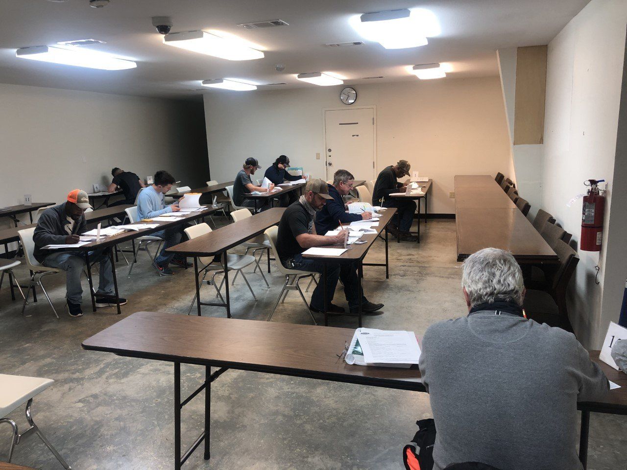 A classroom with people taking a test at long tables. Fluorescent lights illuminate the room.