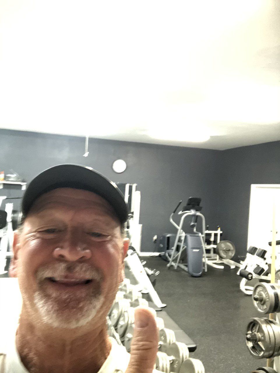 Man in black cap smiles, gives thumbs-up in a gym with weights and exercise equipment.