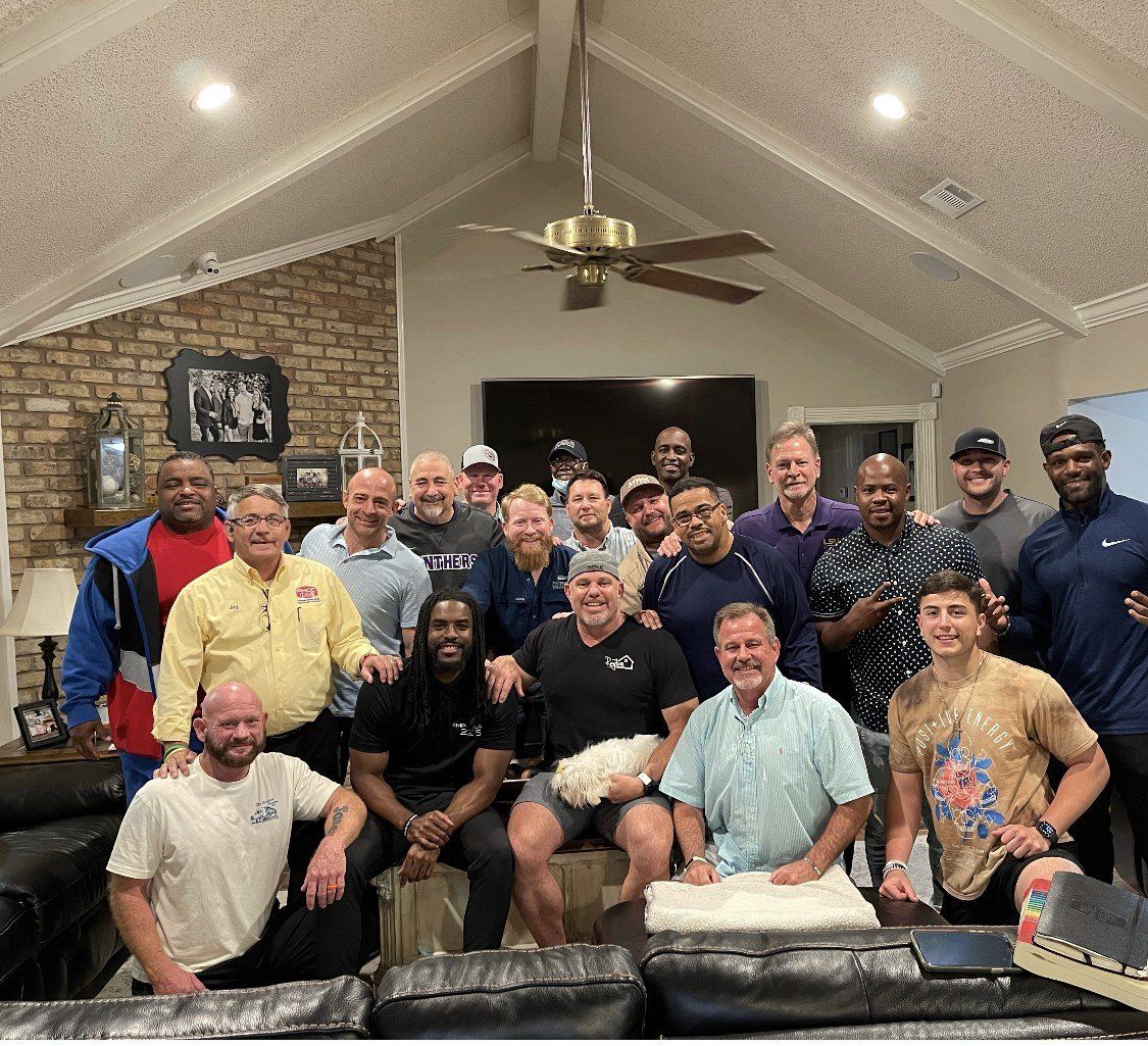 Group of men smiling, gathered in a living room; some seated, some standing.