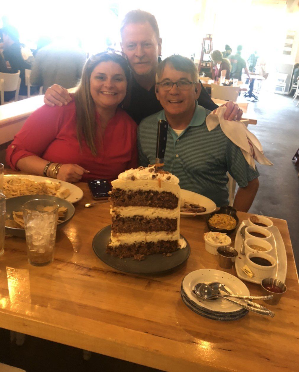 Three people smiling around a large cake with a knife in it at a restaurant.