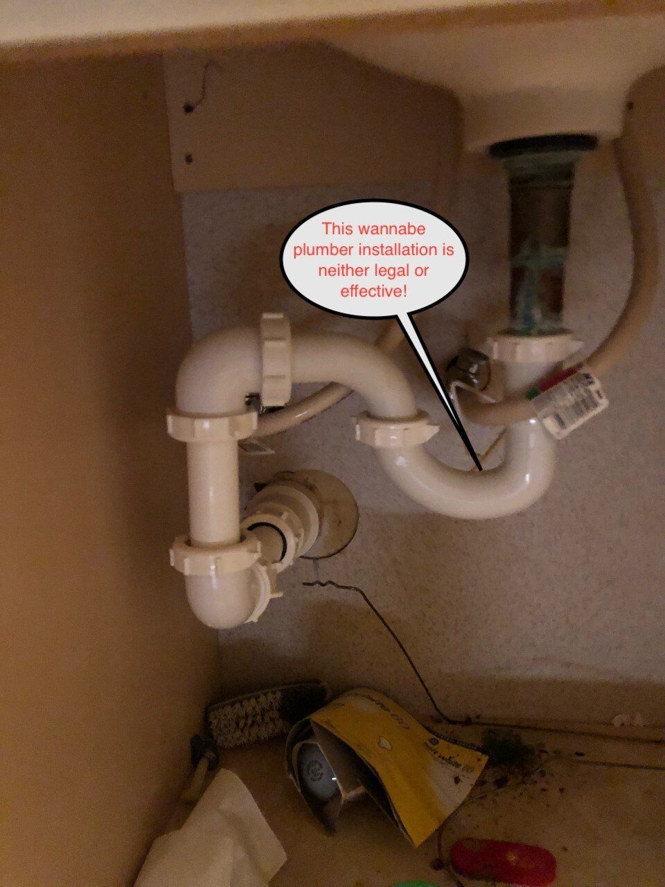 White plastic plumbing under a sink, labeled as an illegal or defective installation.