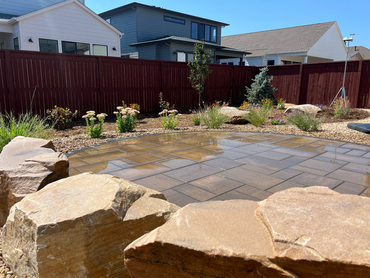 A rocky creek bed with water flows through a vibrant garden, lush green lawn, under a cloudy sky.
